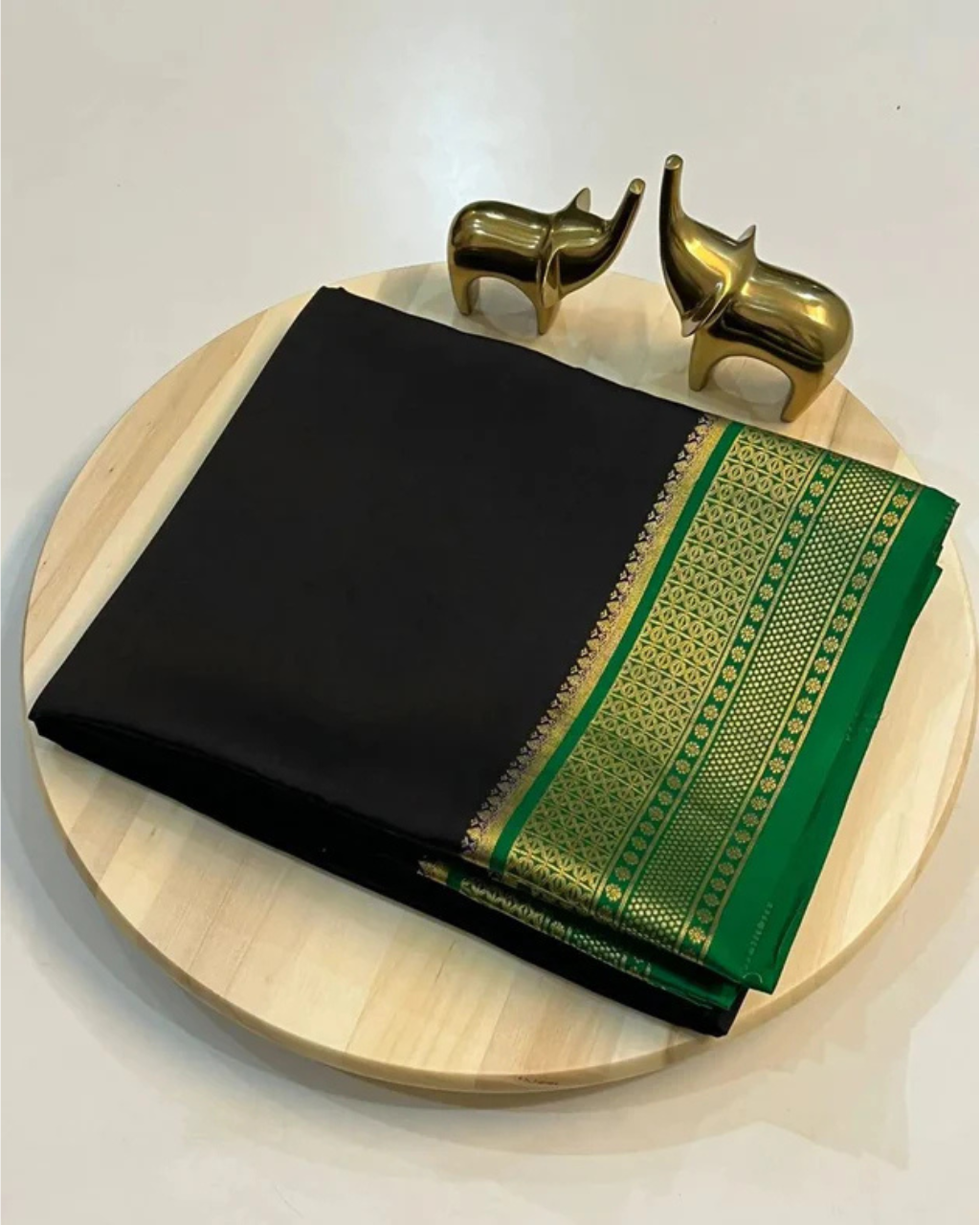 Black and green saree with gold border on a wooden plate, accompanied by two brass elephants.